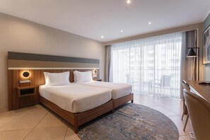 A hotel room with a double bed, bedside tables, a desk, and a large window with sheer curtains. - DoubleTree by Hilton Malta (St. Paul's Bay)