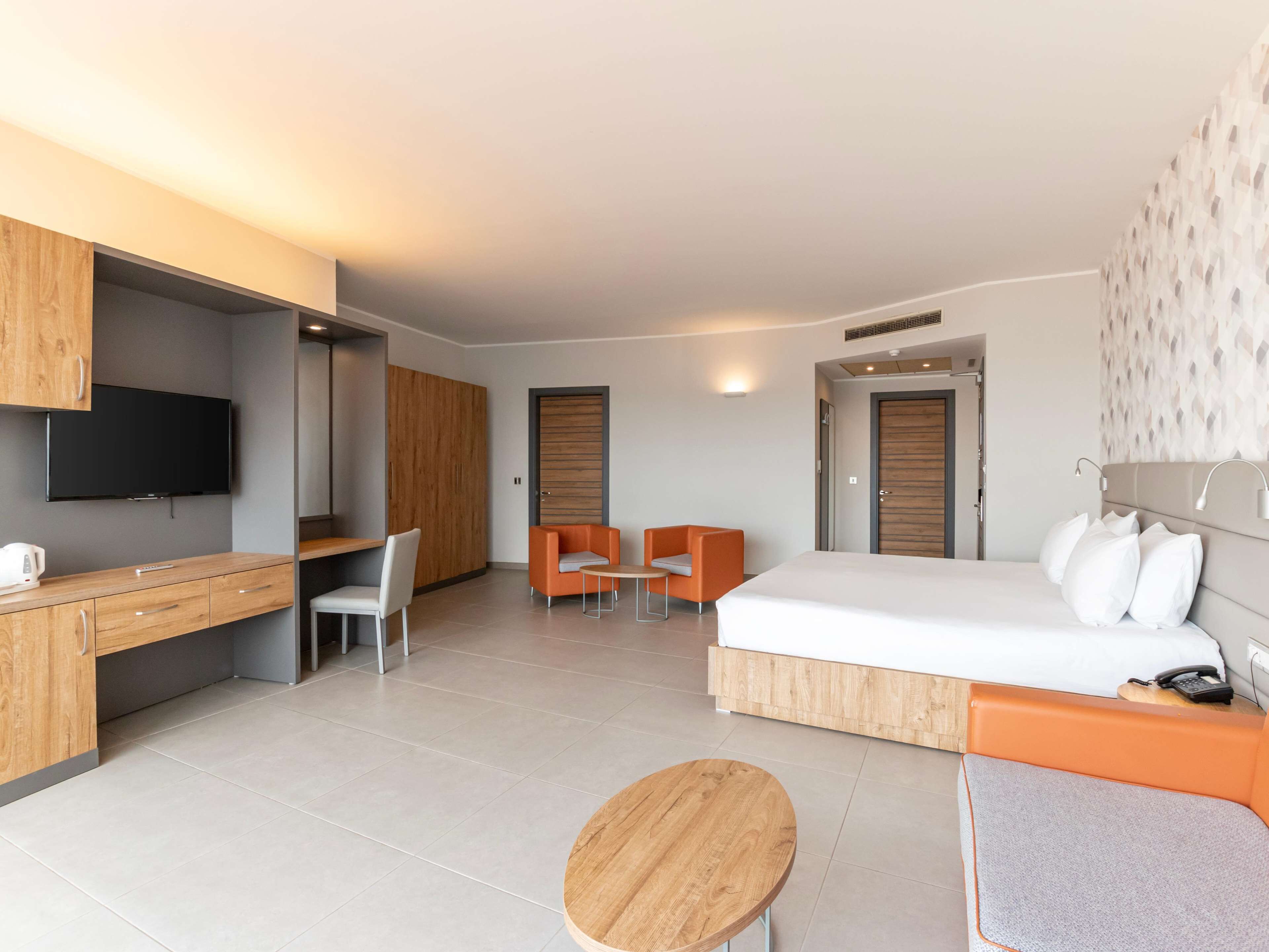 A modern hotel room with a wooden desk, two orange armchairs, a bed, and a TV.