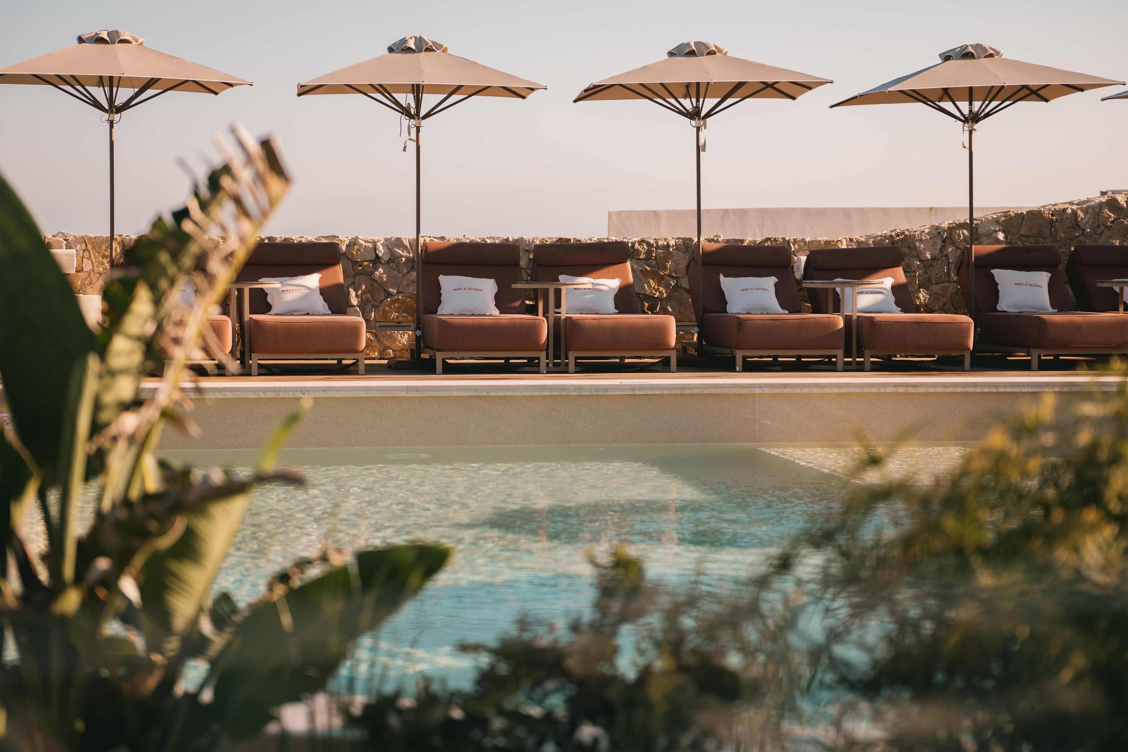A poolside area with sun loungers and umbrellas, a stone wall, and a clear pool.