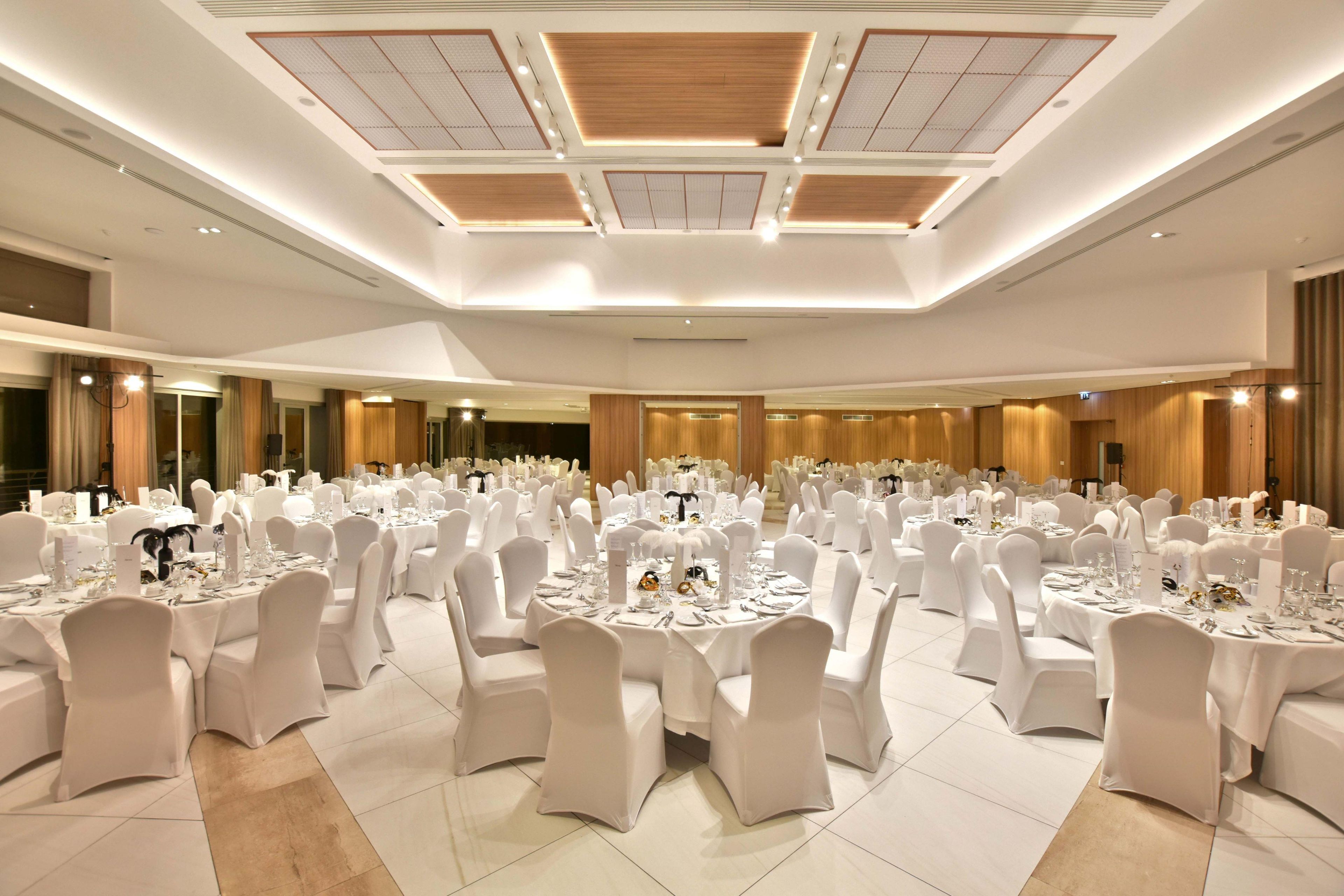 A large banquet hall with round tables, white chairs, and a high ceiling with recessed lighting.