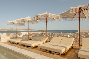 A poolside area with lounge chairs and white macrame umbrellas, overlooking a body of water and rocky landscape.