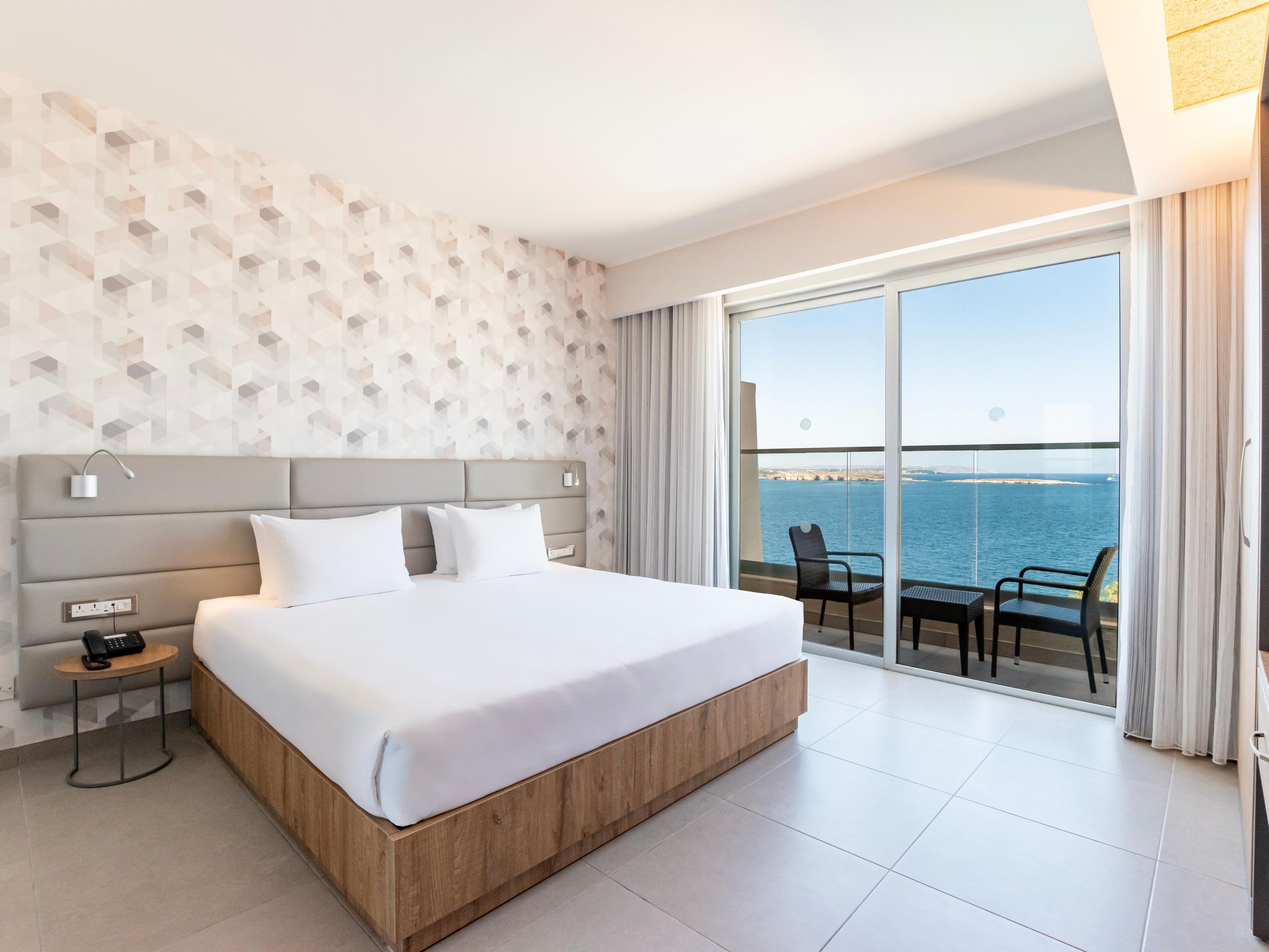 Habitación de hotel con una cama grande, una mesita de noche con teléfono, una pared con diseños geométricos y un balcón con vista al mar.