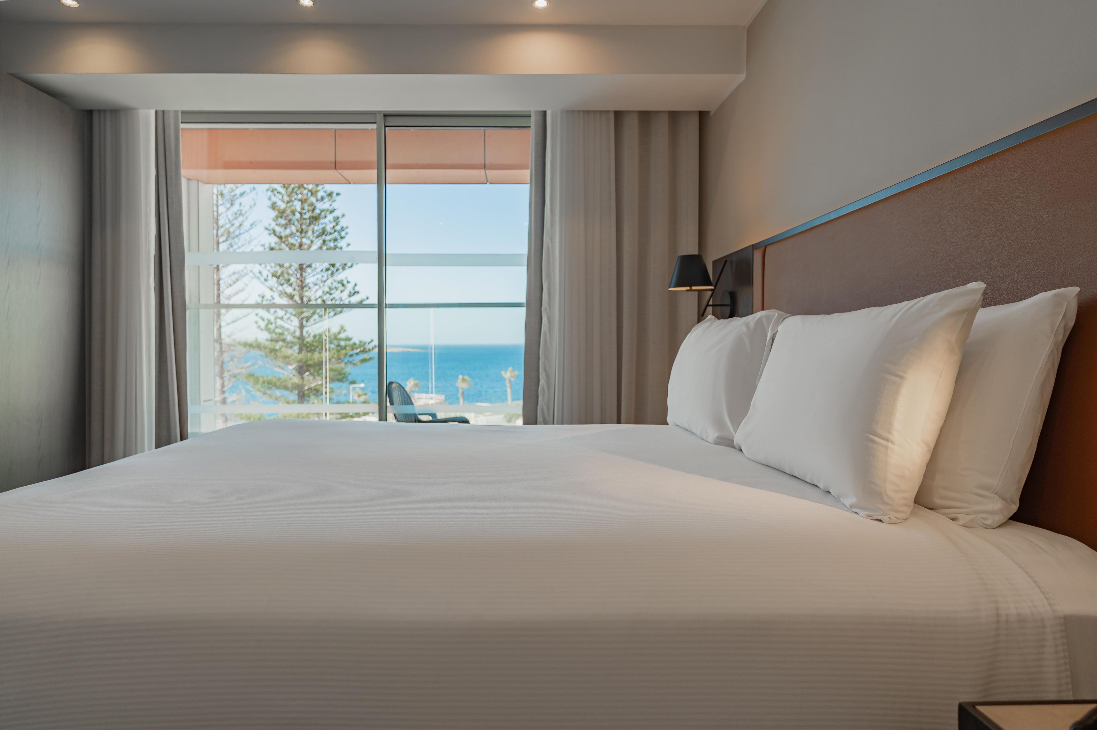 A hotel room with a large bed, a view of the beach, and a chair by the window.