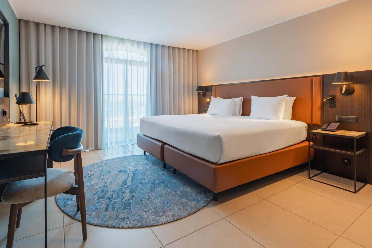 Una habitación de hotel moderna con una cama grande, un escritorio con una silla, una lámpara de escritorio, una mesita de noche con un teléfono y una ventana con cortinas.