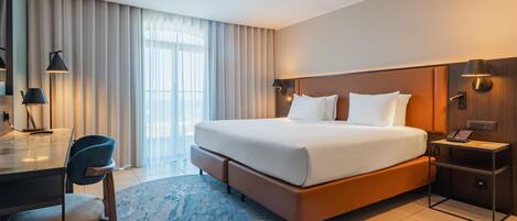 A modern hotel room with a large bed, a desk with a chair, a desk lamp, a nightstand with a phone, and a window with curtains.