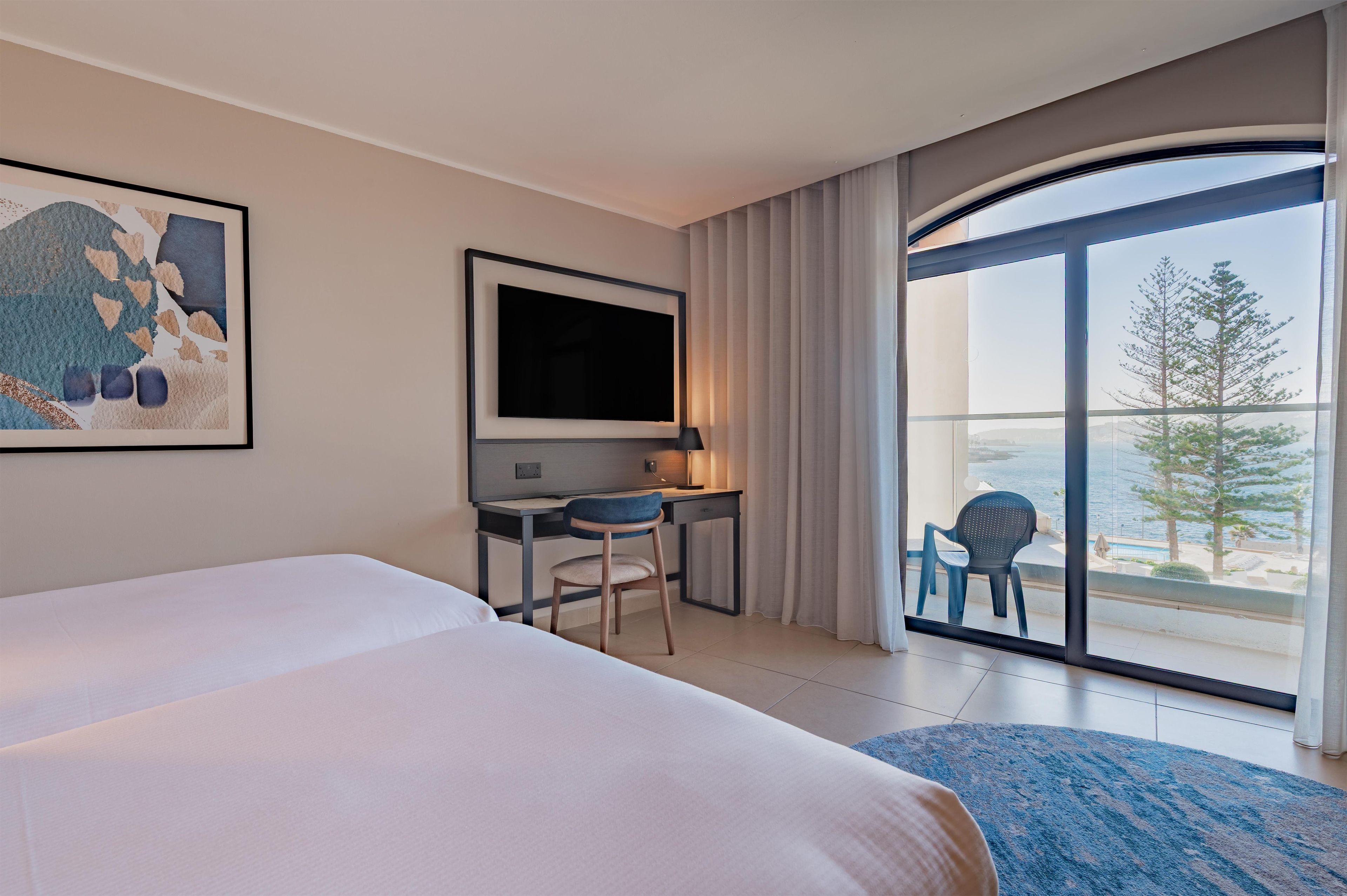 A hotel room with a bed, desk, chair, TV, and a balcony with a view of the sea.