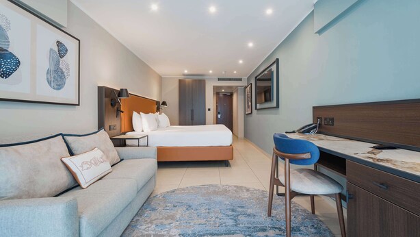 A modern hotel room with a bed, a desk, a chair, a sofa, and artwork on the walls. - DoubleTree by Hilton Malta (St. Paul's Bay)