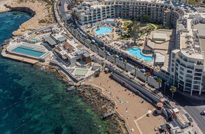 An aerial view of a resort with multiple swimming pools, a beach area, and a large hotel complex. - DoubleTree by Hilton Malta (St. Paul's Bay)