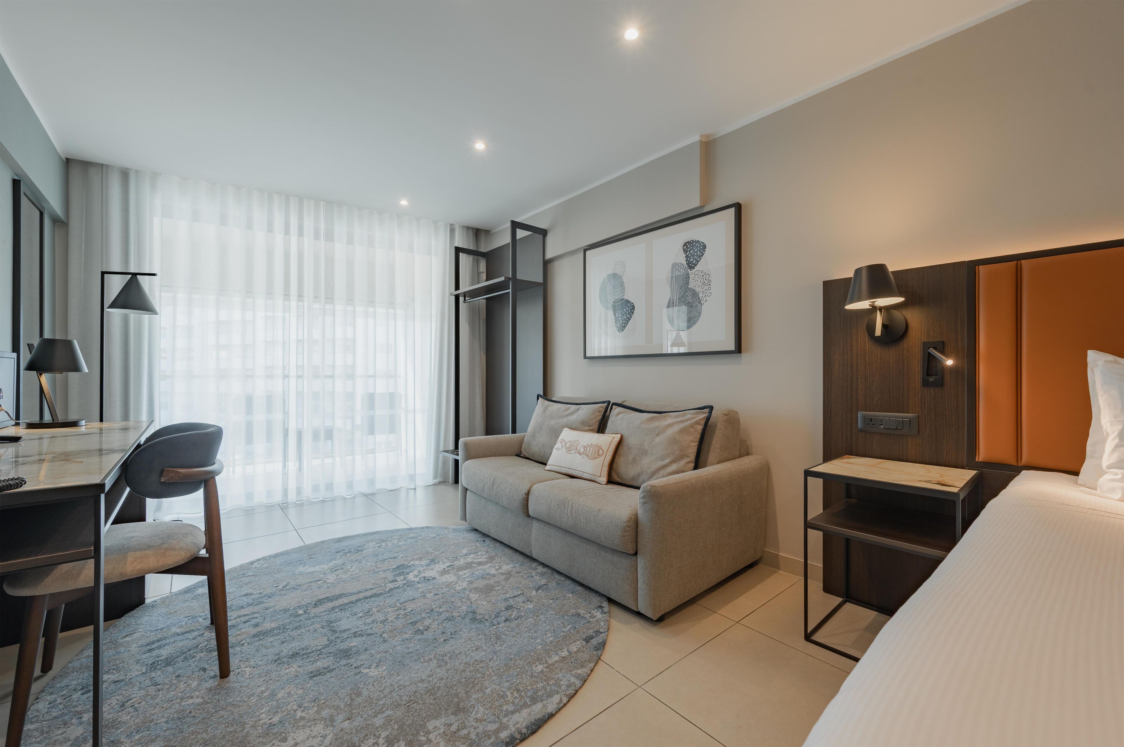 A modern hotel room with a sofa, a desk, a bed, a small table, and a wall-mounted TV.
