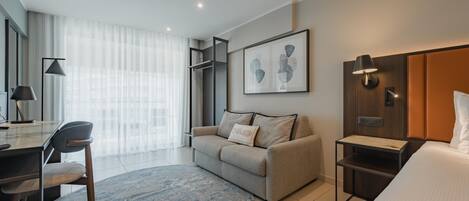 A modern hotel room with a sofa, a desk, a bed, a small table, and a wall-mounted TV.