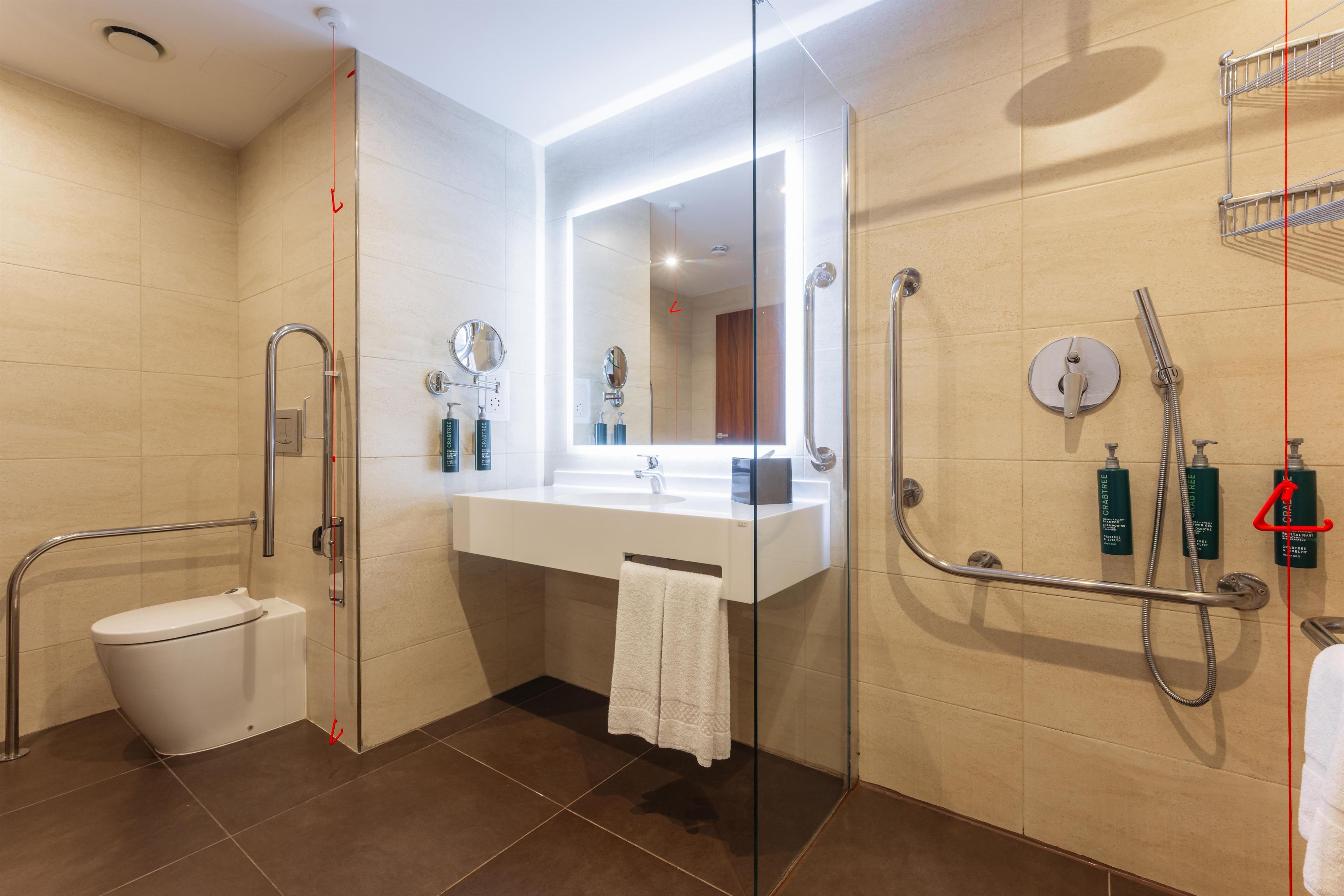Une salle de bain moderne avec une douche à parois de verre, des toilettes blanches, un lavabo avec un miroir et des barres d’appui.