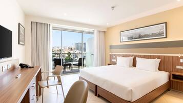 Ein Hotelzimmer mit einem großen Bett, einem Schreibtisch mit Stuhl, einem Fernseher und Blick auf die Stadt durch die Fenster.