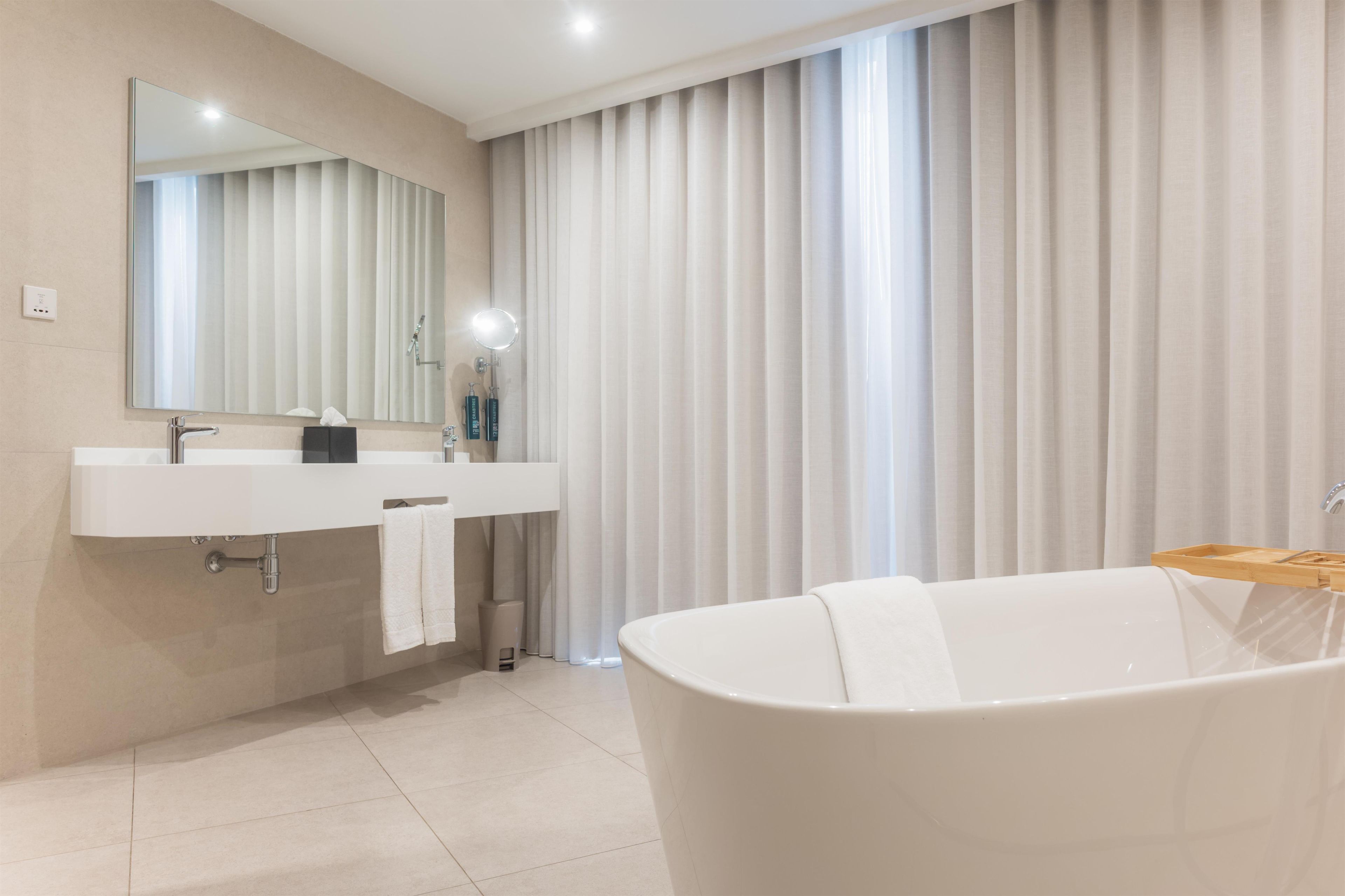 Een moderne badkamer met een groot bad, een wastafel met spiegel en een raam met verticale zonwering.