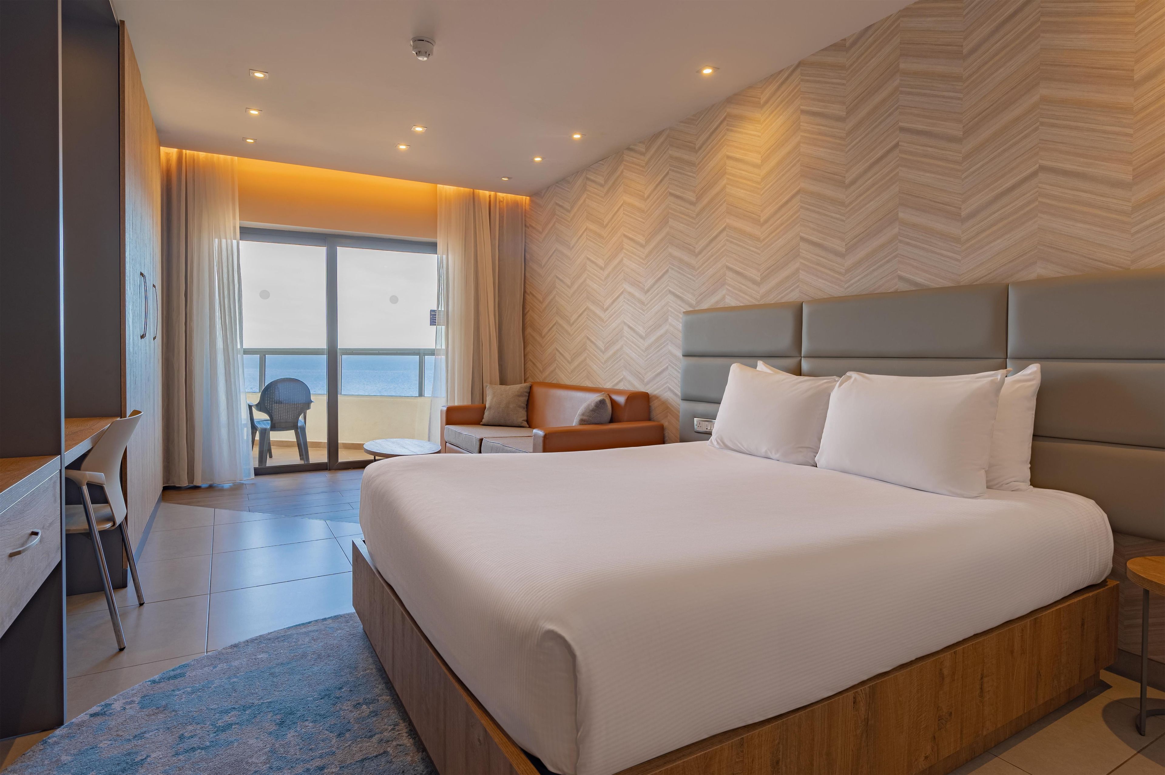 A modern hotel room with a large bed, a seating area with a sofa and armchairs, a desk, and a view through a glass door to the outside.