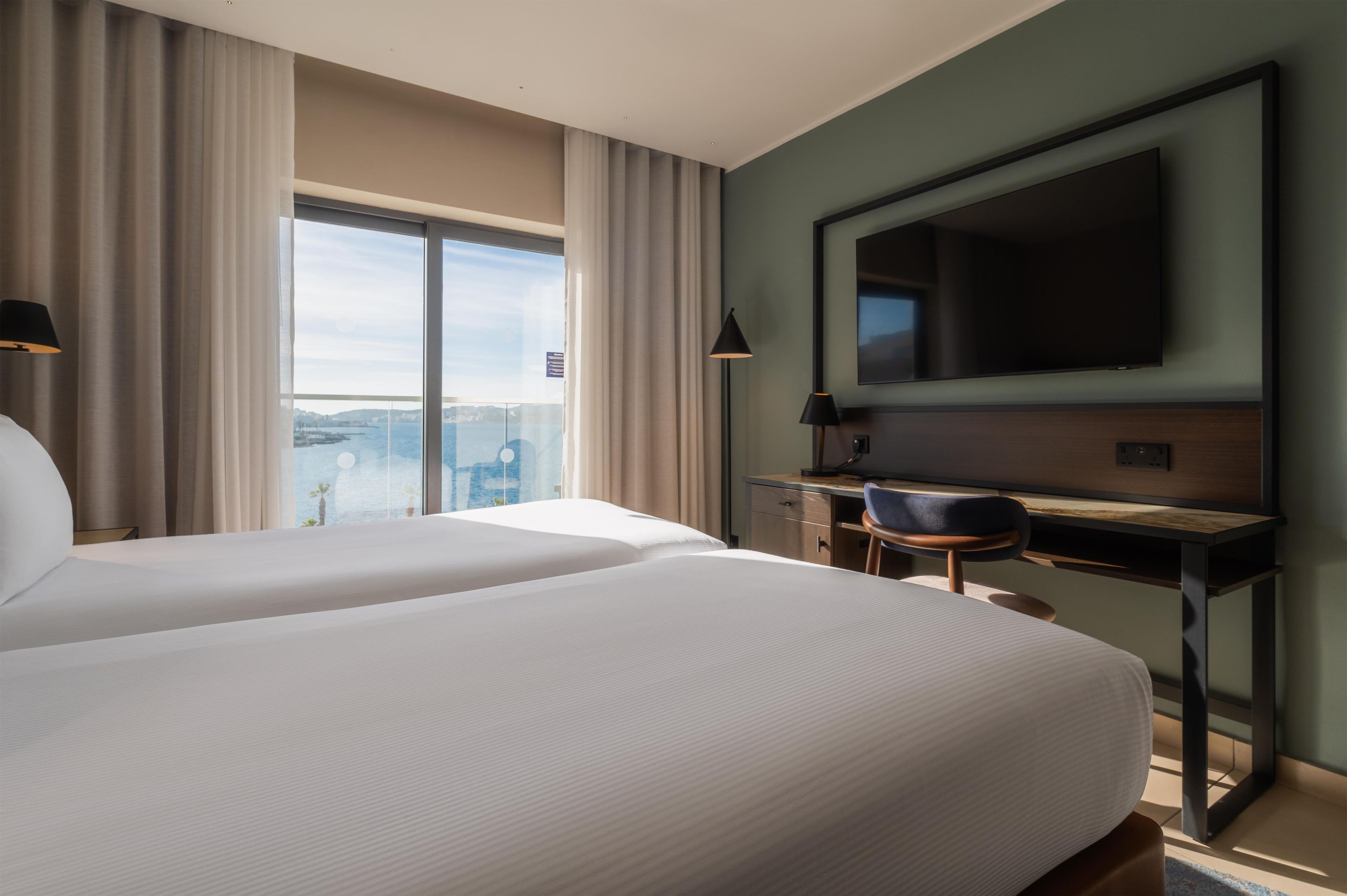 A hotel room with two beds, a desk, a chair, a television, and a view of the city.