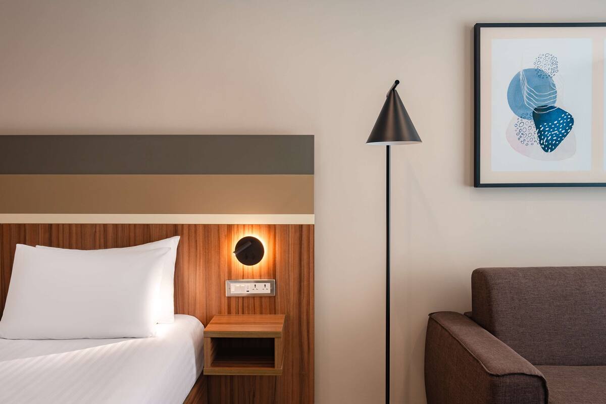 Una habitación de hotel con una cama, una mesita de noche con una lámpara, una obra de arte enmarcada en la pared y un sofá.