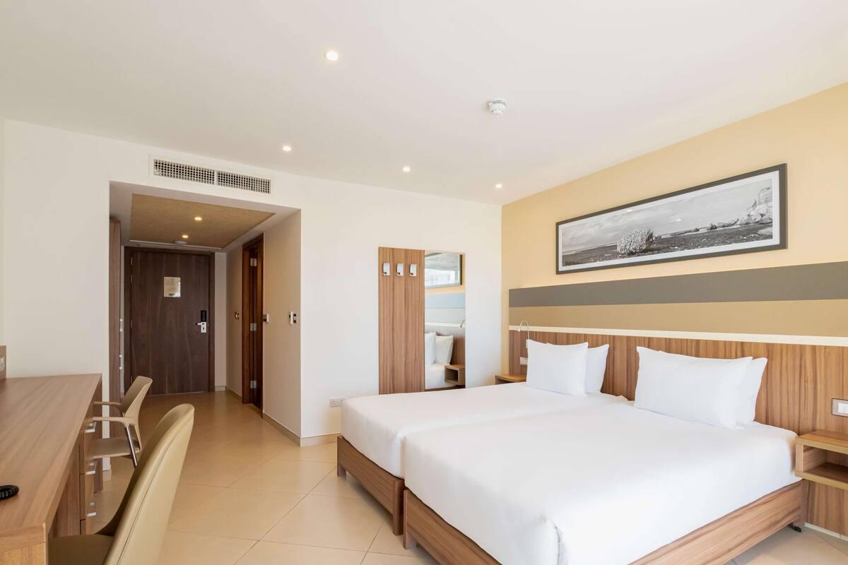 Habitación de hotel con una cama grande, un escritorio y una silla. Hay un espejo en la pared y un cuadro sobre la cama.