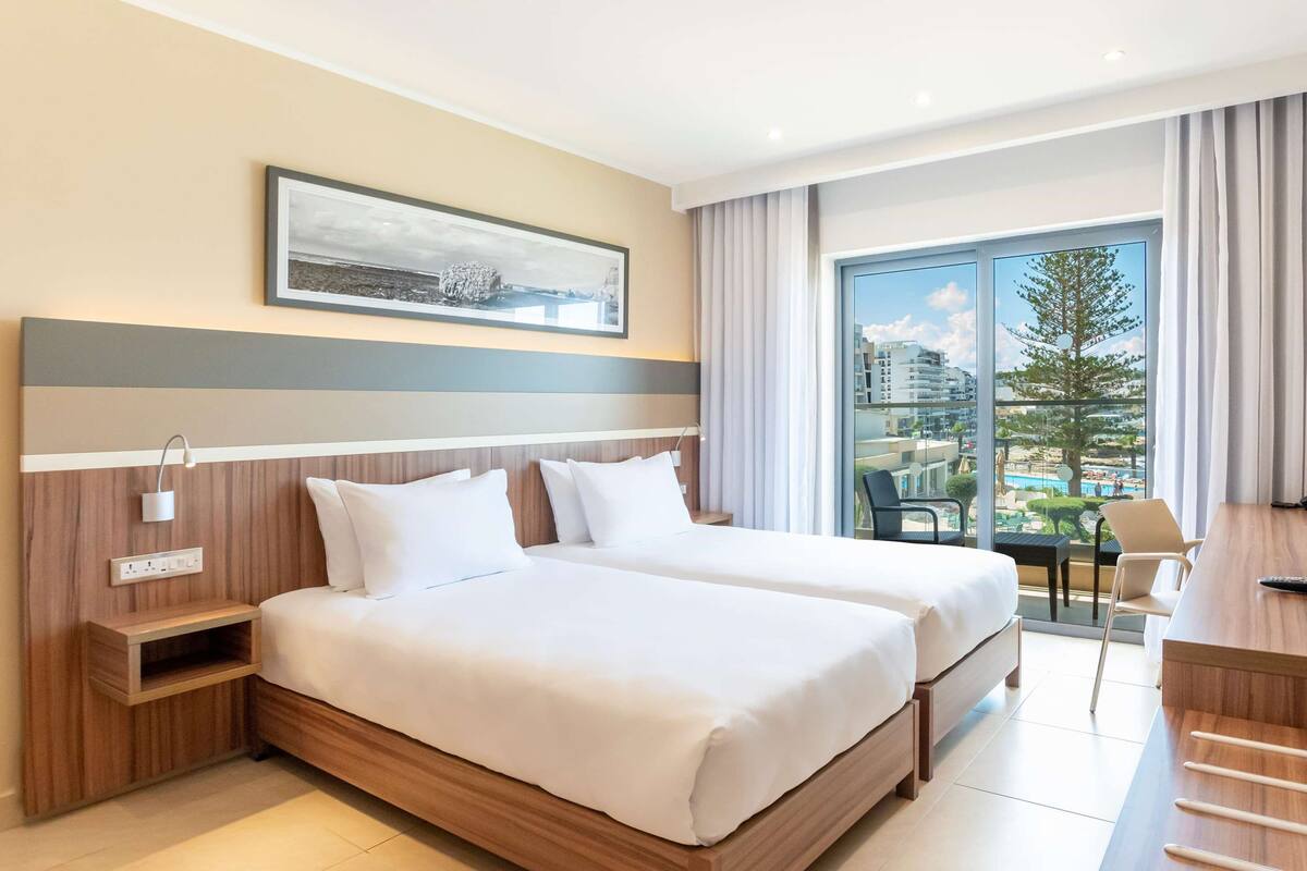 Habitación de hotel con dos camas, un escritorio, un televisor y un ventanal con vistas a edificios y un árbol.