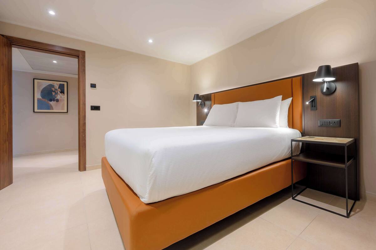 Una habitación de hotel moderna con una cama grande, una mesita de noche con lámpara y una puerta que da a otra habitación.