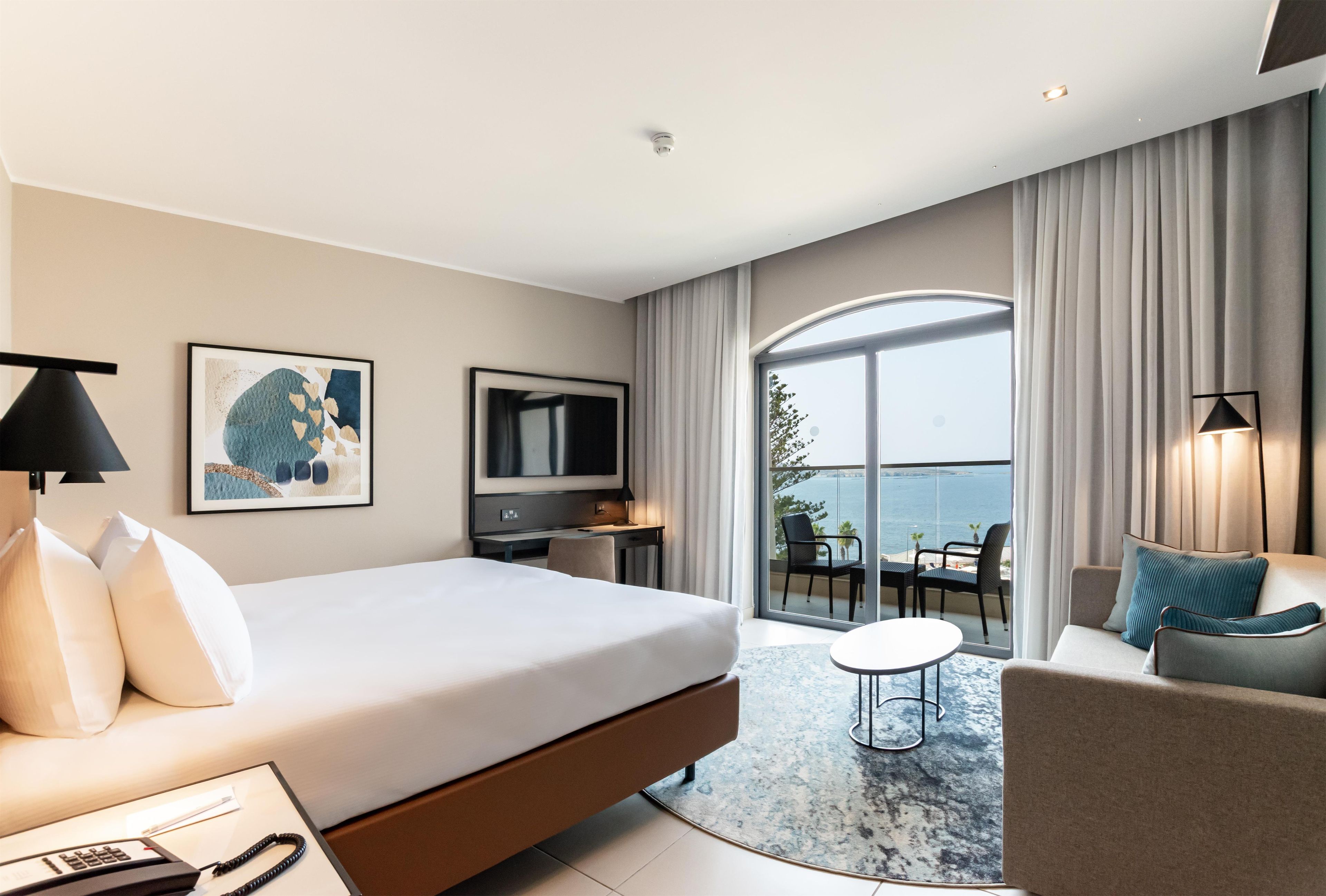 Une chambre d’hôtel comprenant un lit, un bureau, une télévision, un balcon avec vue sur la mer, ainsi qu’un coin salon avec une table et des chaises.