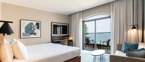 A hotel room with a bed, a desk, a TV, a balcony with a view of the sea, and a seating area with a table and chairs.