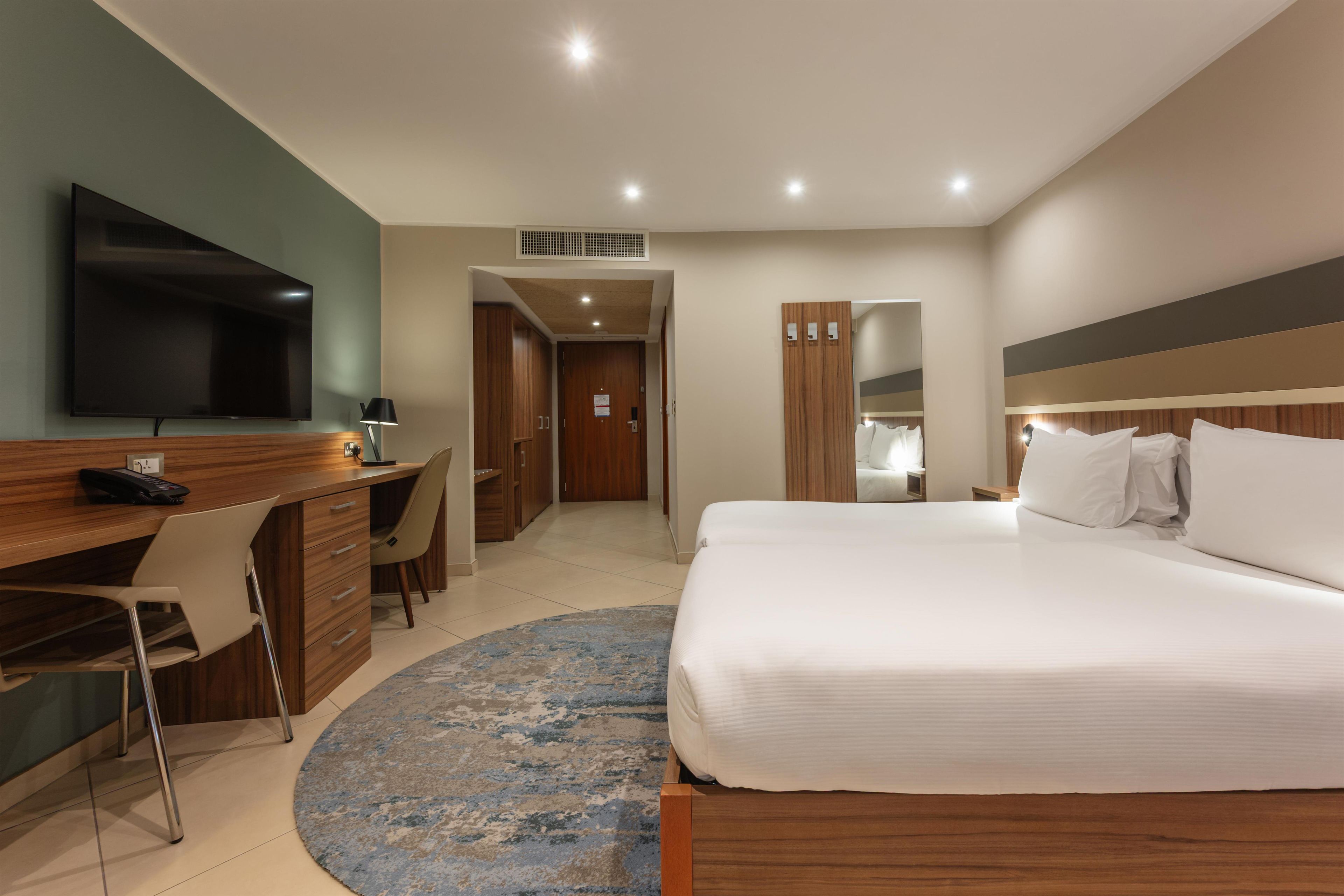 A hotel room with a bed, a desk with a chair, a television, and a bathroom visible through an open door.