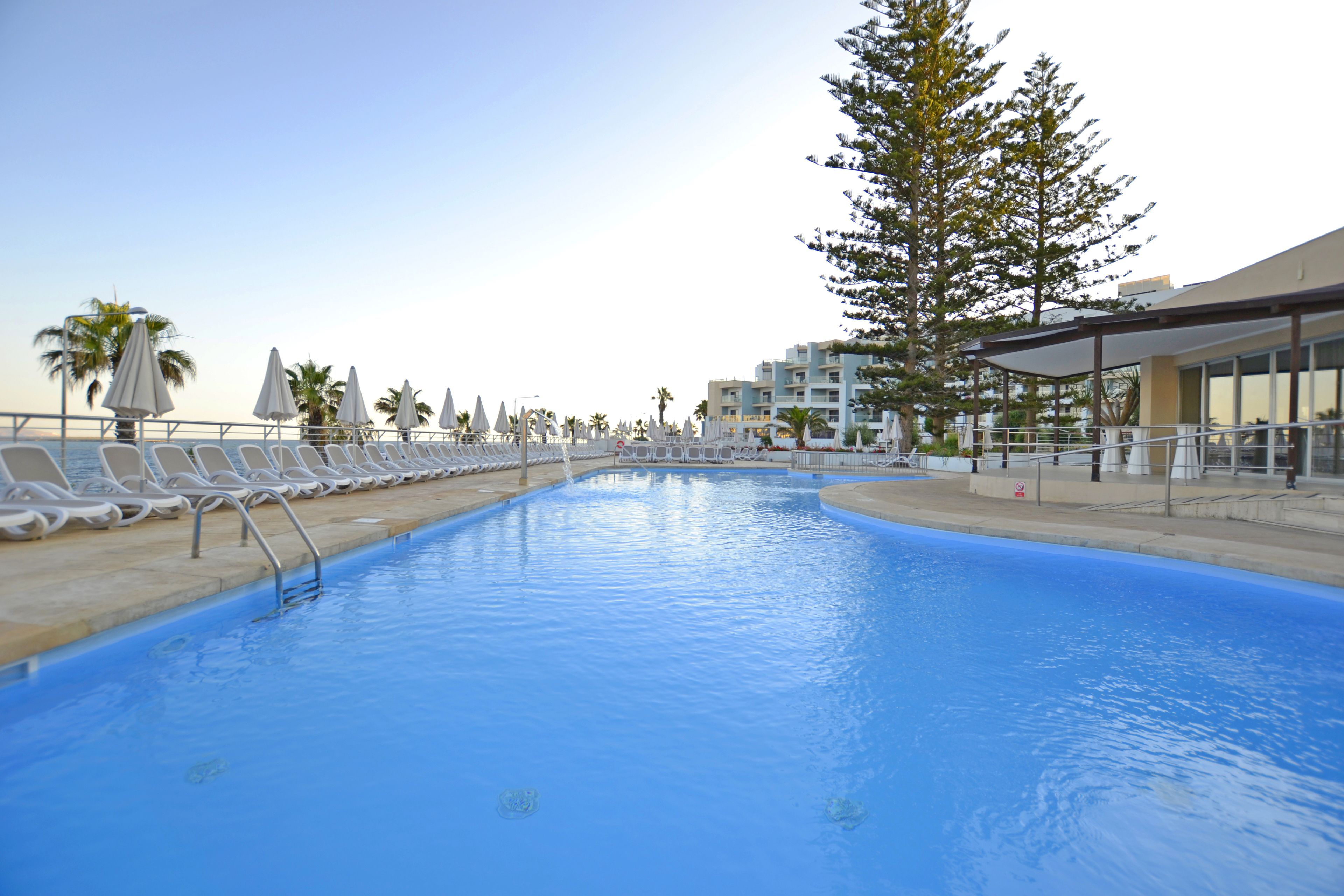 Una piscina con sillas de descanso y sombrillas, rodeada de edificios y árboles.