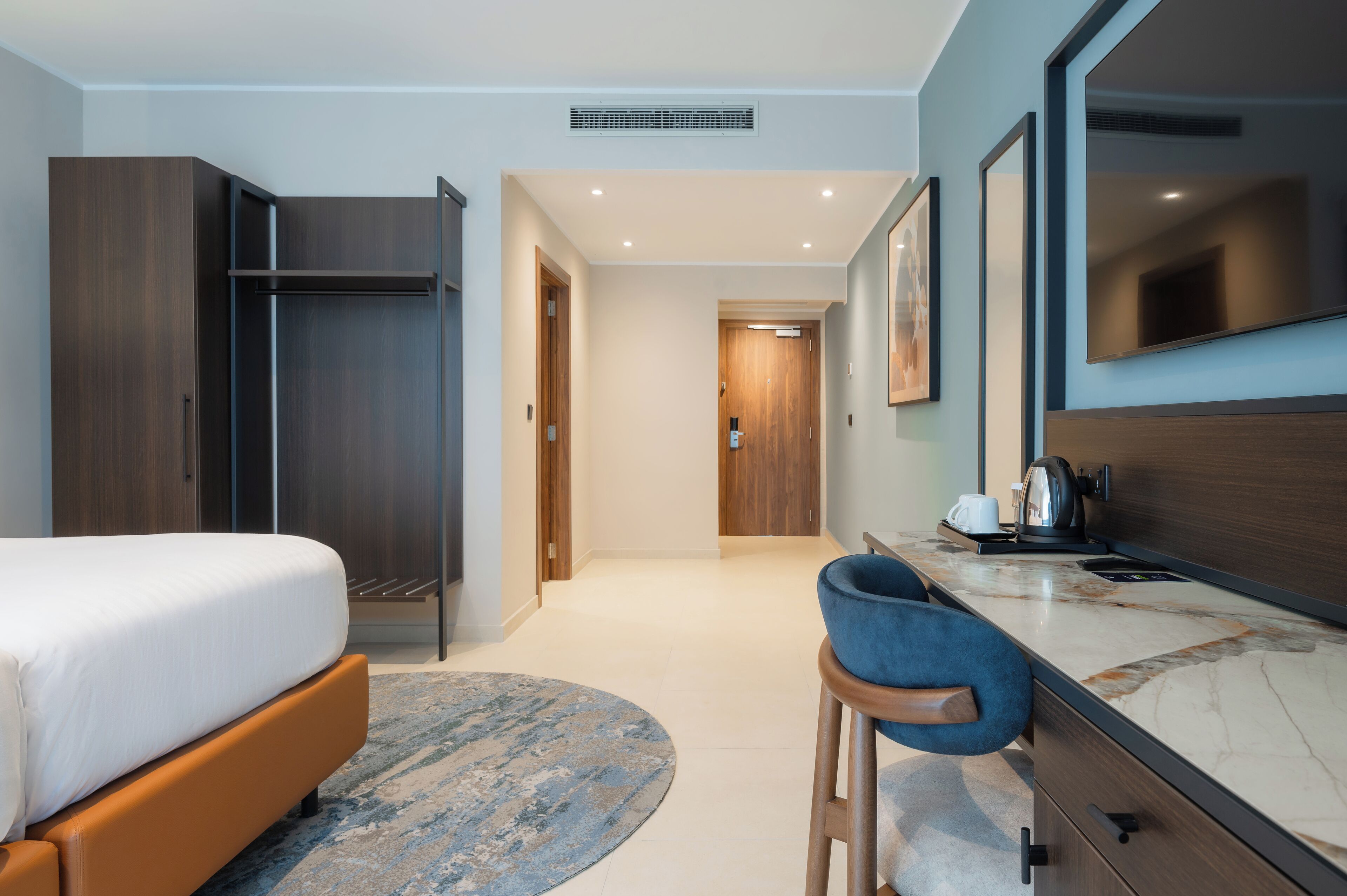 Et moderne hotelværelse med seng, skrivebord med stol, stort spejl og garderobeskab.