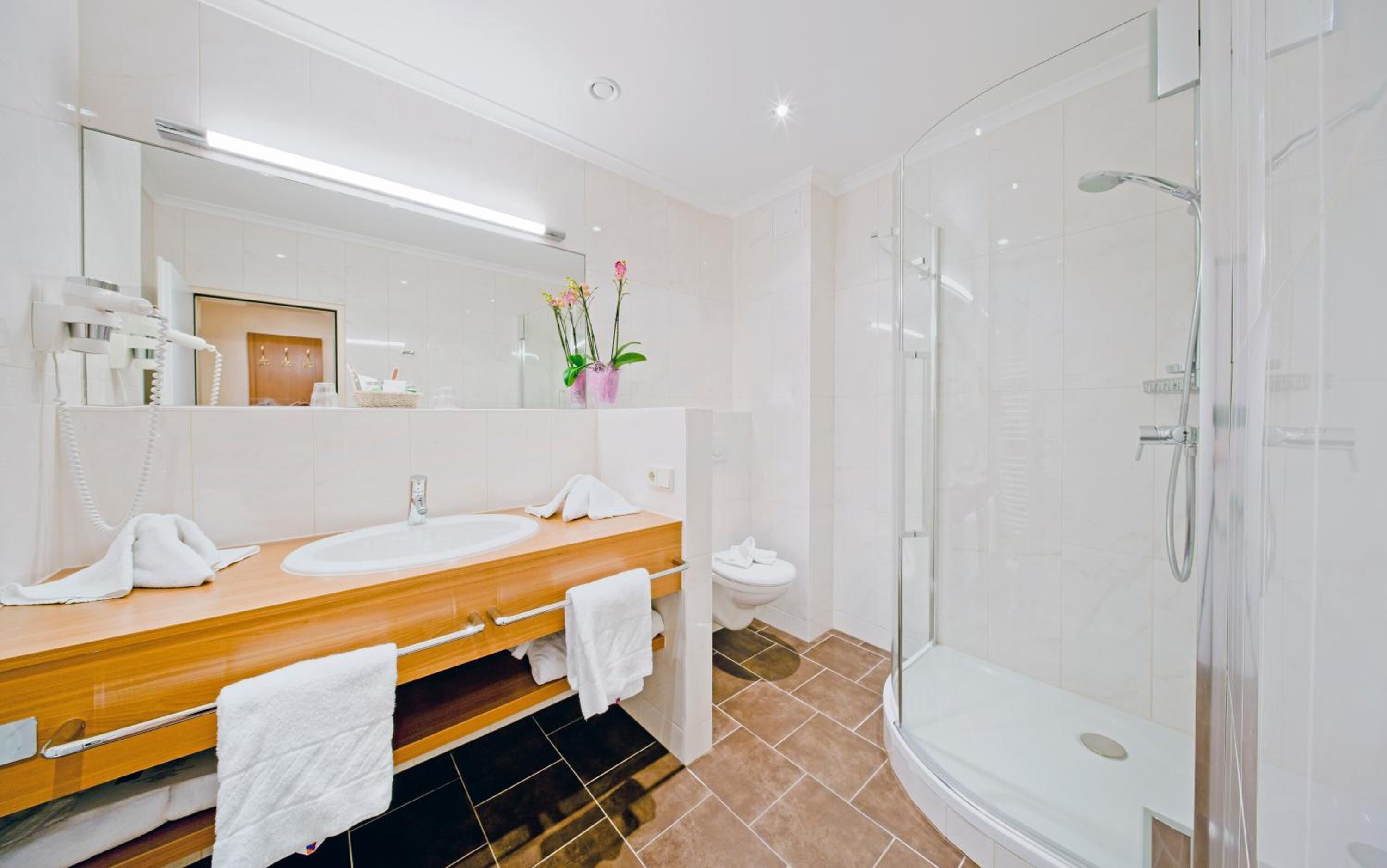 A modern bathroom with a wooden vanity, two sinks, a glass shower enclosure, and a mirror with a vase of flowers.