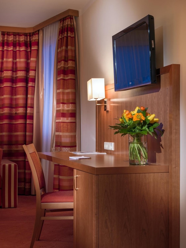 A hotel room with a wooden desk, a chair, a television mounted on the wall, a lamp, and a vase with flowers.