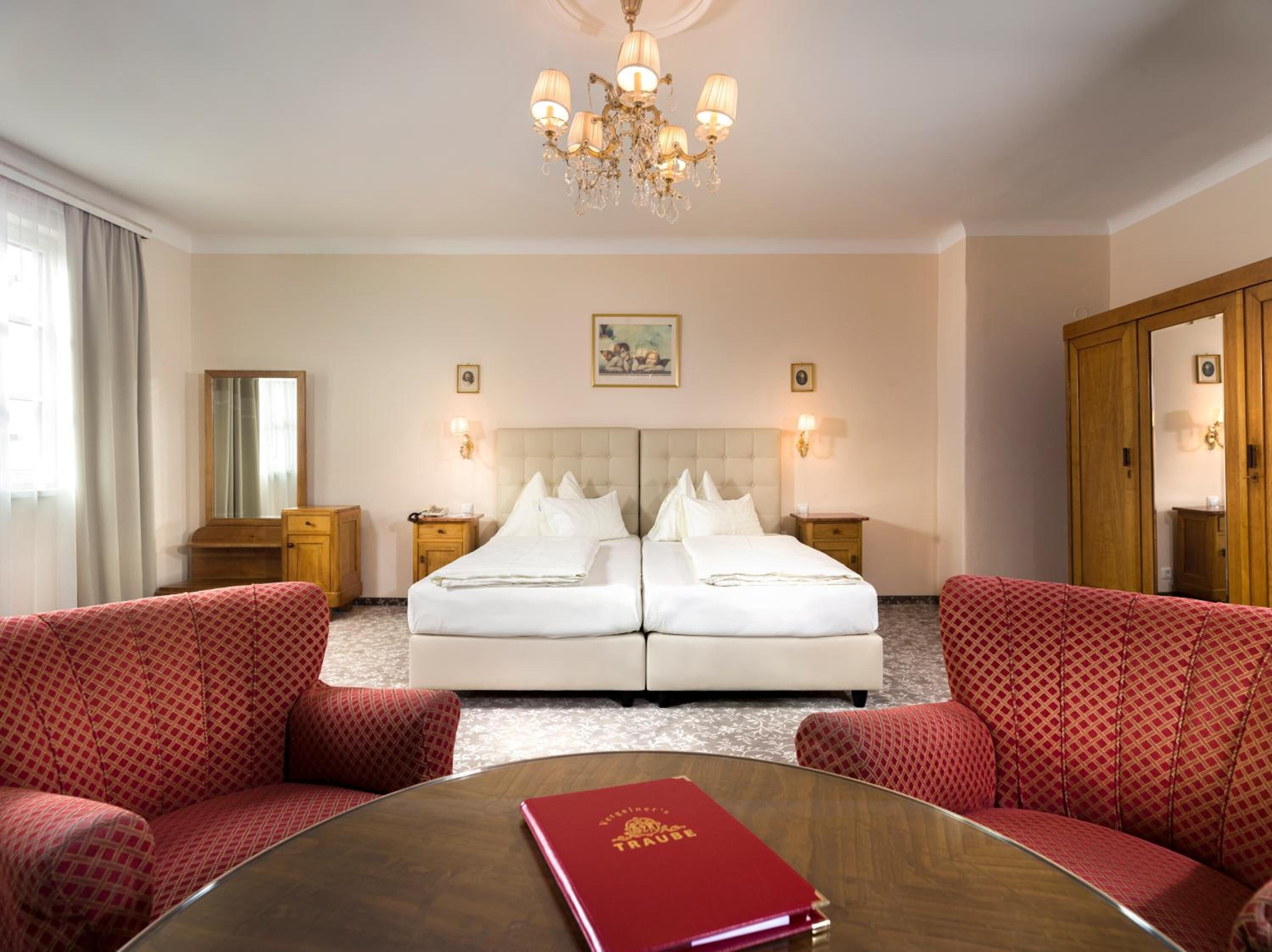 A hotel room with a double bed, two bedside tables, a chandelier, a mirror, a red armchair, a round table with a red book, and a large wardrobe.