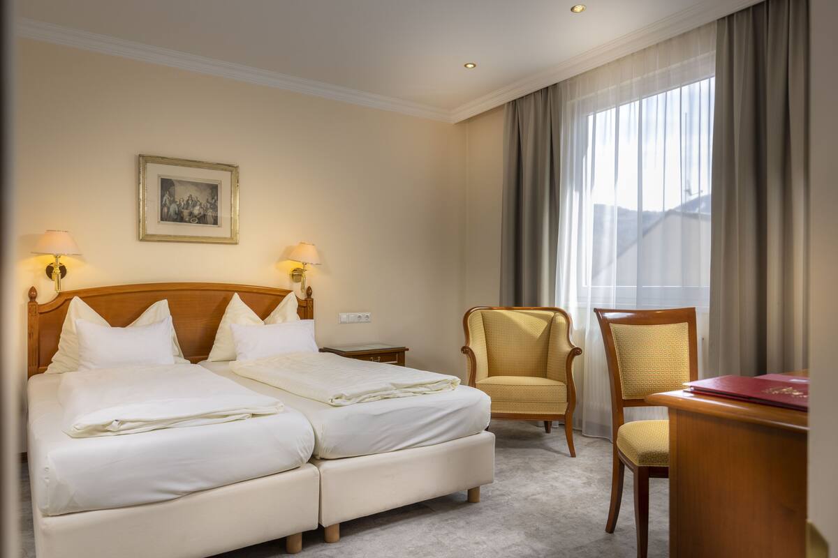 Habitación de hotel con cama doble, dos mesitas de noche, dos lámparas, una silla, un escritorio y una ventana con cortinas.
