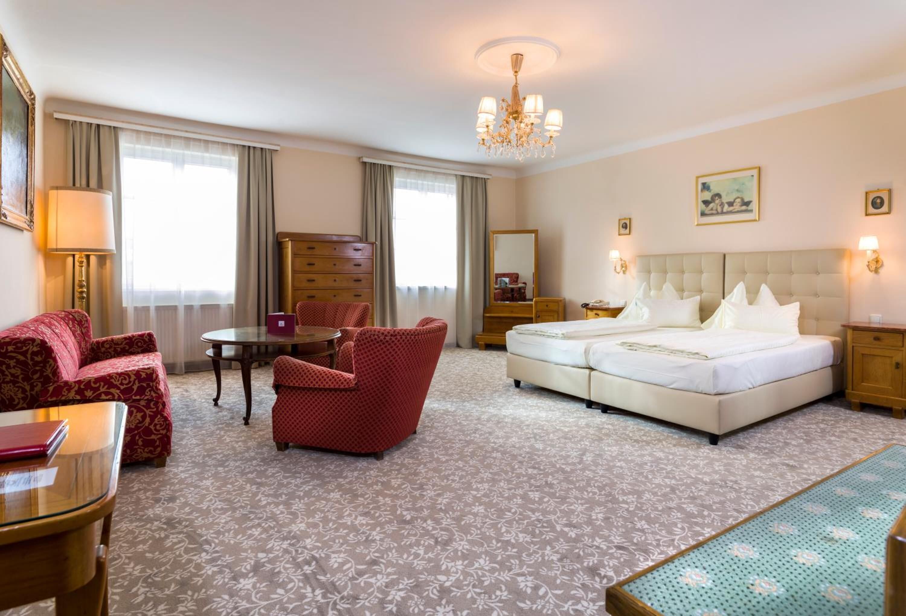 A hotel room with a bed, two red armchairs, a small table, a lamp, a dresser, and a mirror.
