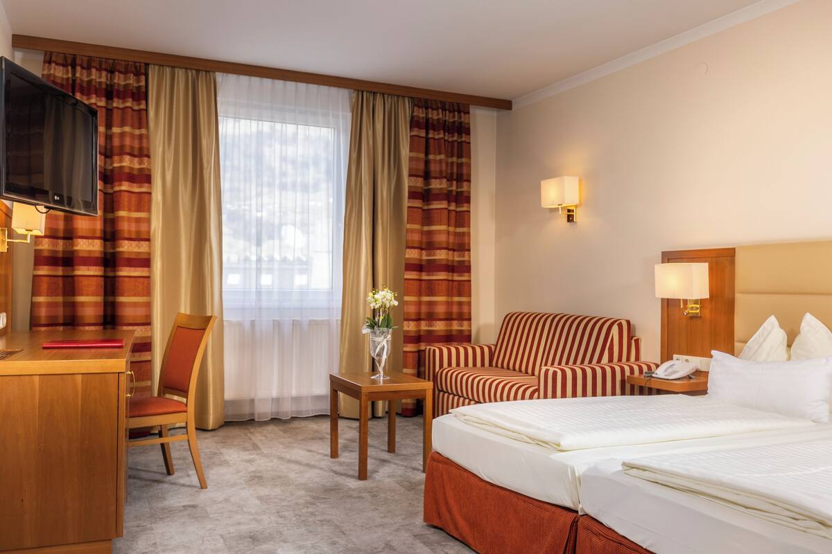Habitación de hotel con cama, escritorio, silla, sofá, televisor y ventana con cortinas.