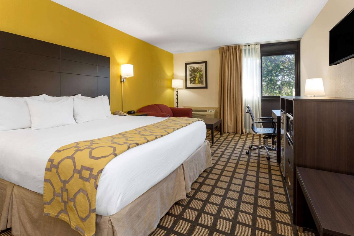 Ein Hotelzimmer mit einem großen Bett, einem Schreibtisch, einem Sessel, einem Fernseher und einem Fenster mit Vorhängen.