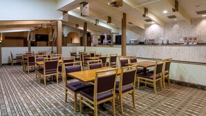 Un restaurante con mesas y sillas de madera, una zona de cocina y una barra con alimentos.