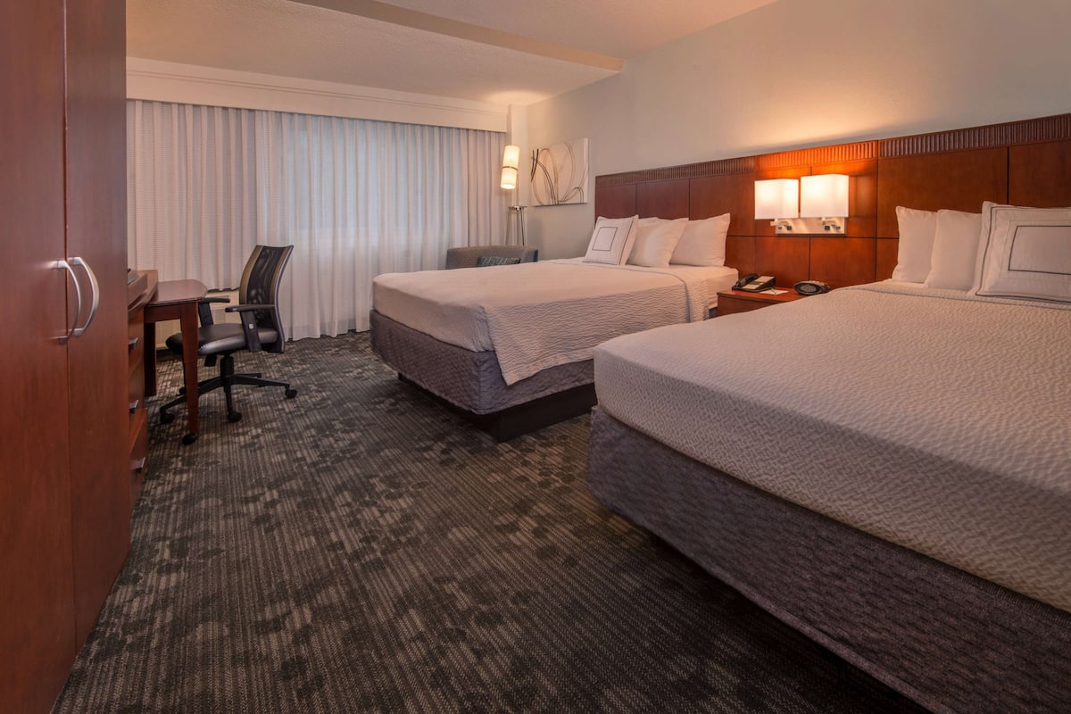 A hotel room with two beds, a desk, a chair, and a large window with curtains.