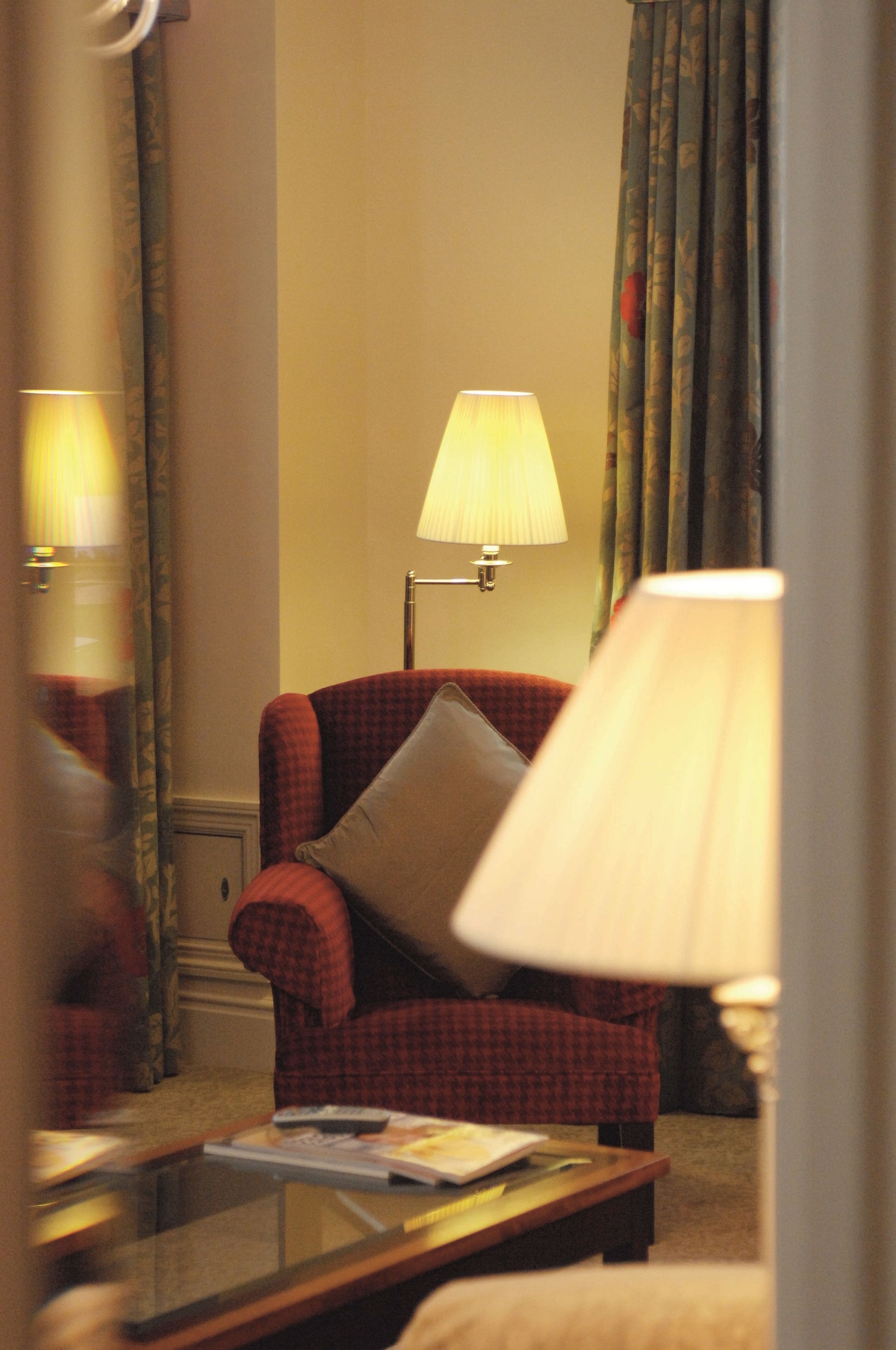 A hotel room with a checkered armchair, a standing lamp, a glass coffee table, and a mirror reflecting the room's decor.
