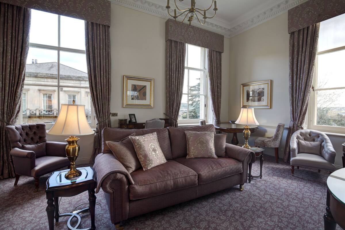 Une pièce avec un canapé marron, deux fauteuils, une table et une lampe. Il y a deux fenêtres avec des rideaux et un lustre au plafond.