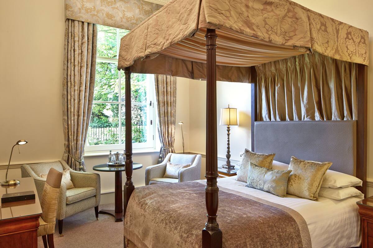 Un lit à baldaquin à quatre poteaux, doté d’une tête de lit, de deux fauteuils, d’une petite table et d’une lampe, dans une pièce bien éclairée grâce à de grandes fenêtres et à des rideaux.