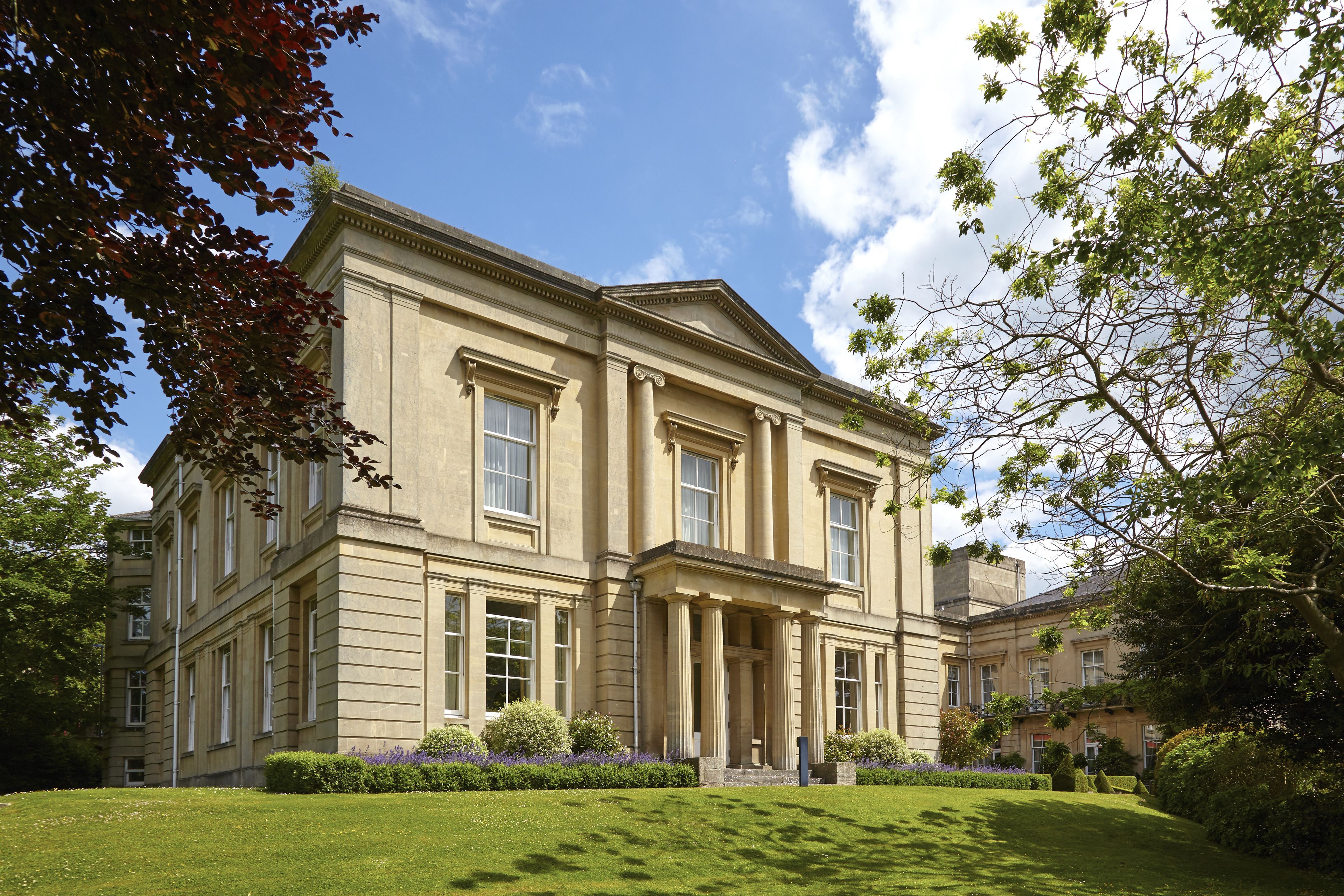 A large, classical building with symmetrical architecture, featuring a central entrance with columns, surrounded by well-maintained lawns and mature trees.
