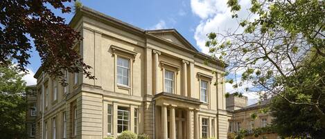 A large, classical building with symmetrical architecture, featuring a central entrance with columns, surrounded by well-maintained lawns and mature trees.
