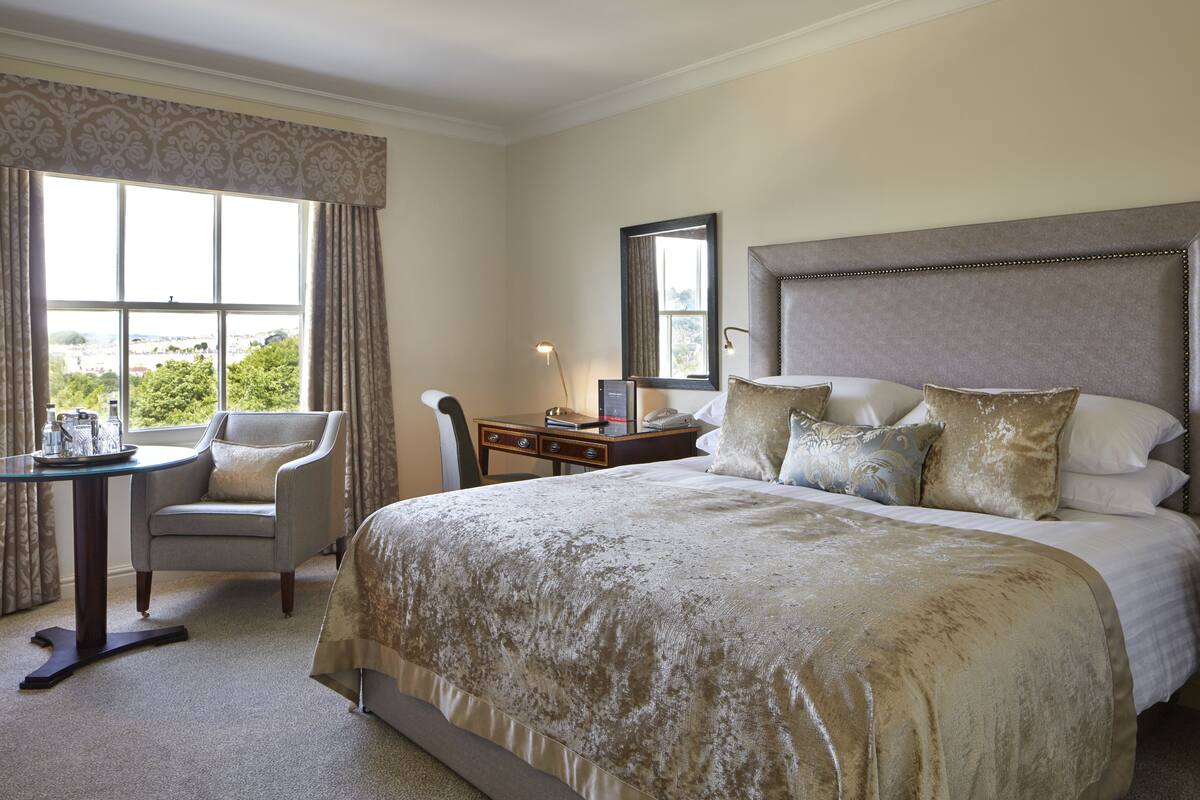 Une chambre d’hôtel avec un grand lit, un bureau avec une chaise, une fenêtre avec des rideaux et une vue sur la verdure.