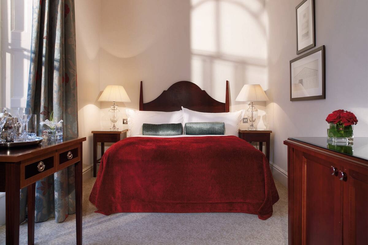 Une chambre d’hôtel avec un grand lit, deux tables de chevet avec des lampes, une tête de lit en bois, une petite armoire et une fenêtre avec des rideaux.