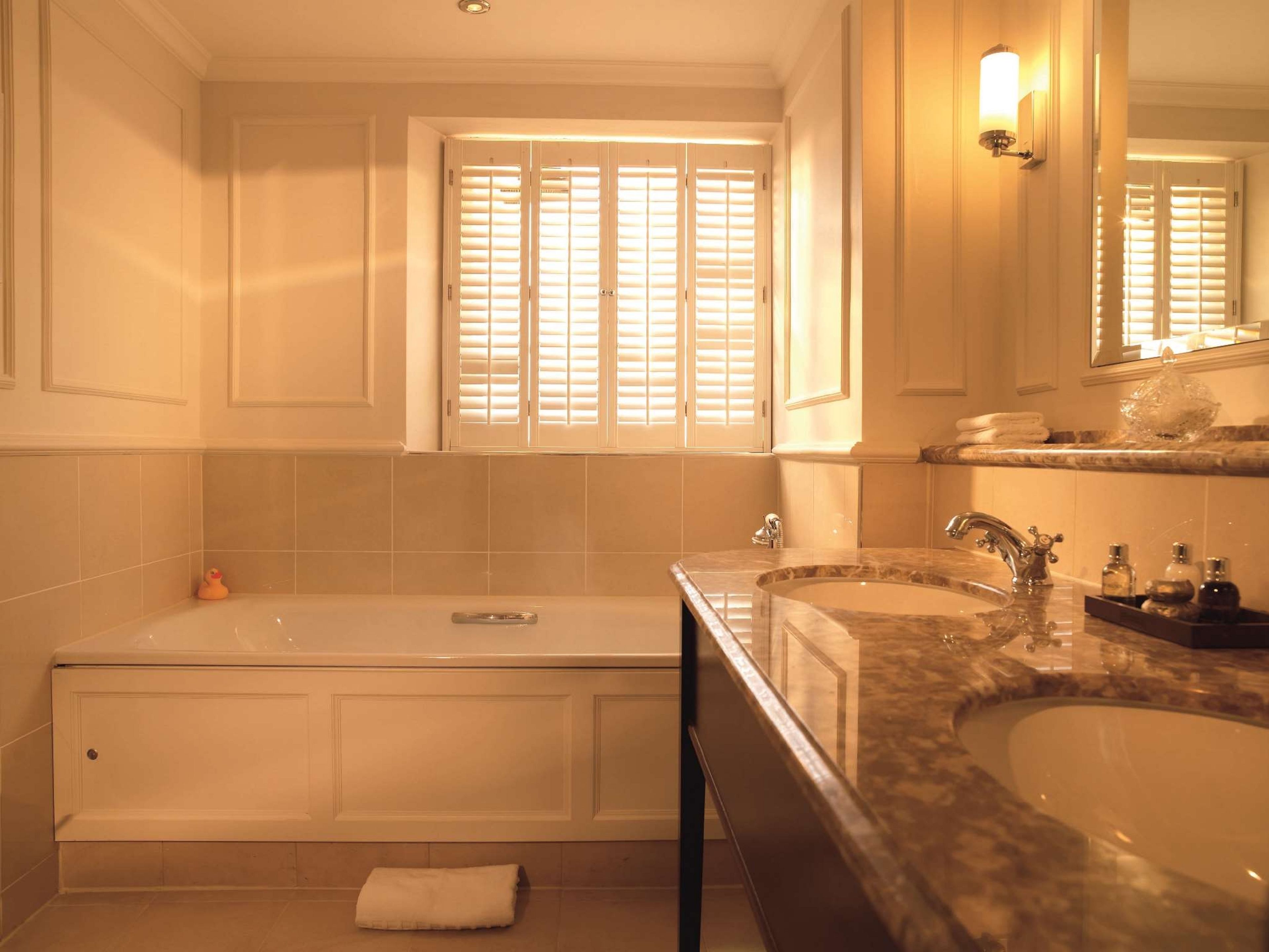 A bathroom with a bathtub, a double sink, and a window with blinds.