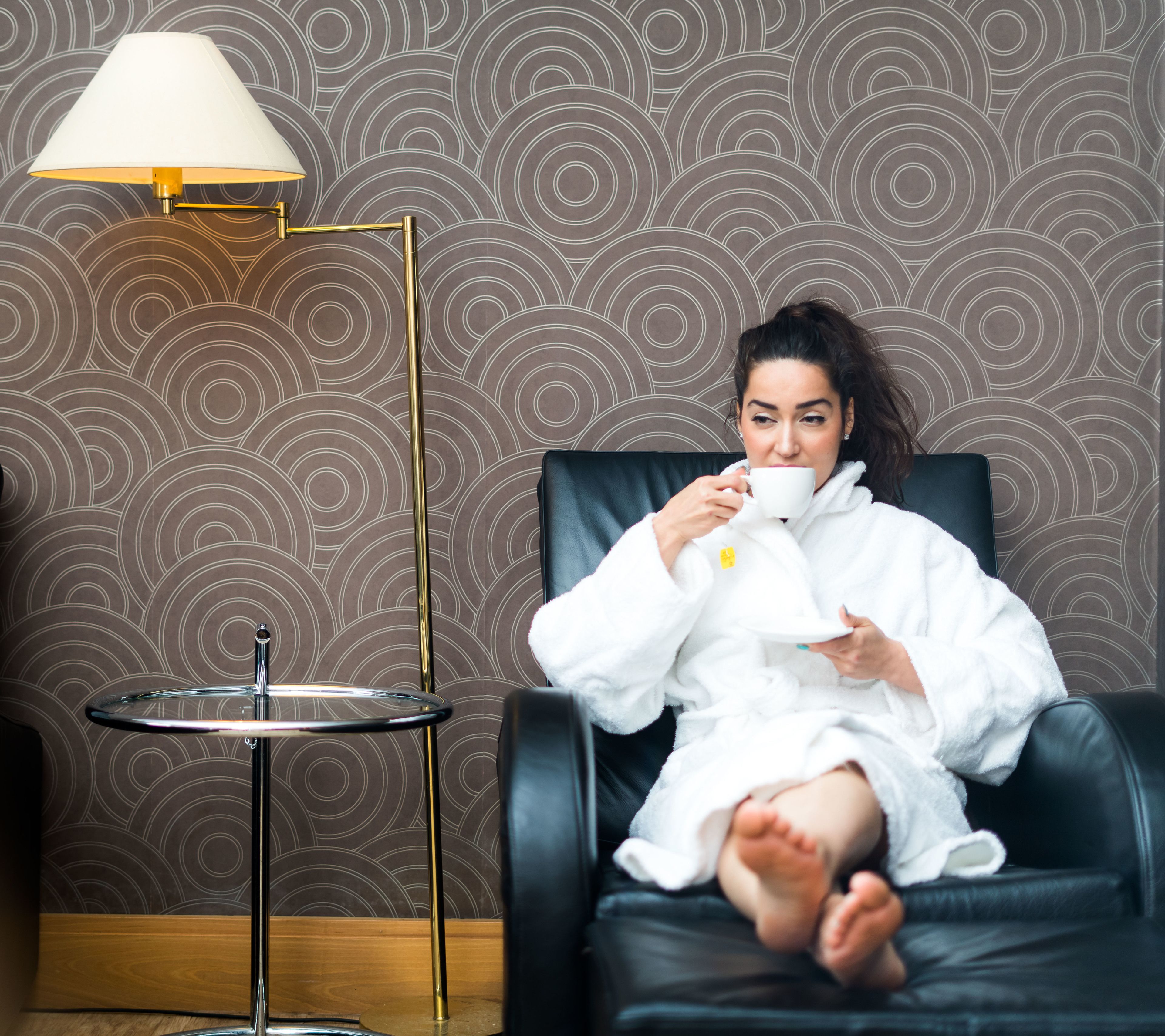 Une personne vêtue d’un peignoir blanc est assise sur un fauteuil en cuir noir. On aperçoit une tasse blanche, une lampe de sol dorée et un papier peint à motifs en arrière-plan.
