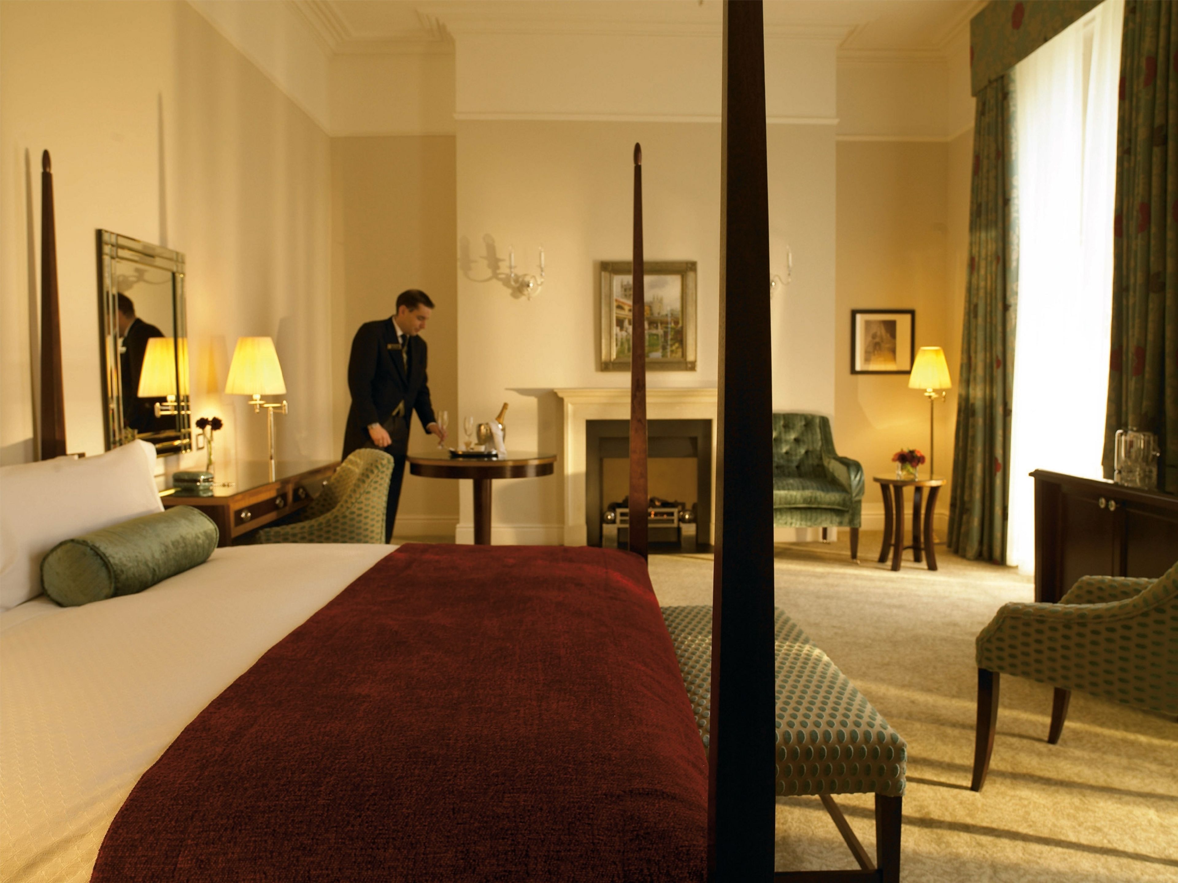Une chambre d’hôtel avec un lit à baldaquin, un coin salon comprenant un fauteuil et une petite table, une cheminée et un miroir au mur.