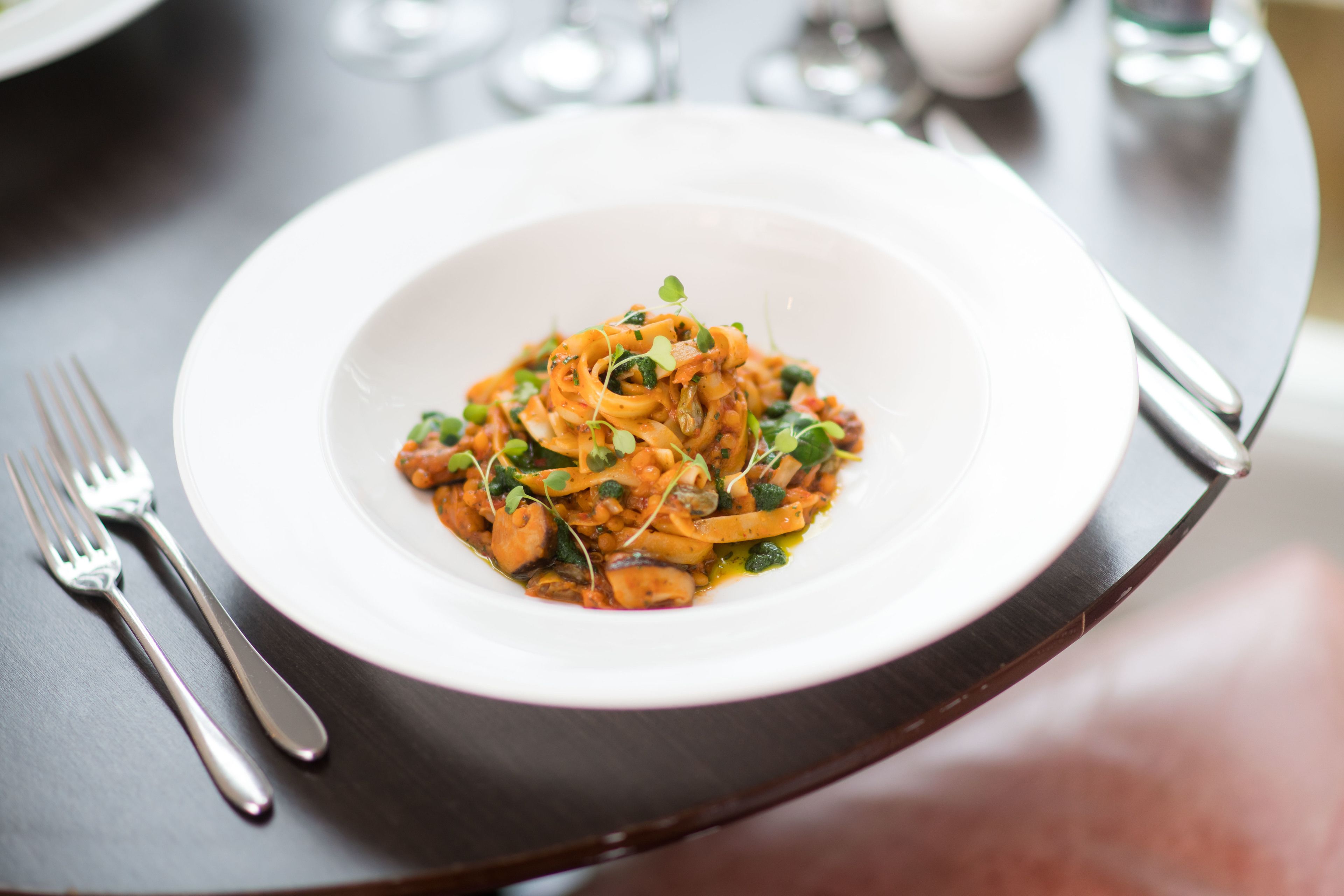 A white plate of pasta with greens and sauce on a dark table.