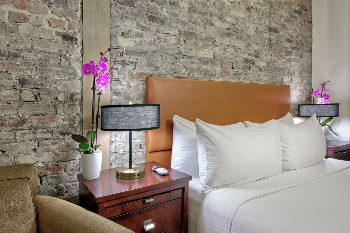 A hotel room with a brick wall, a wooden headboard, a white bed, a nightstand with a lamp, and a potted orchid.