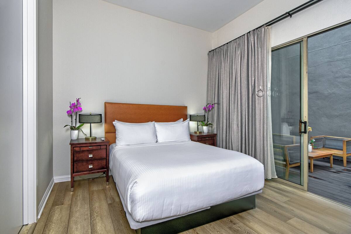 A hotel room with a large bed, wooden floor, a nightstand with a lamp, a small table with a chair, and a sliding glass door leading to an outdoor area.